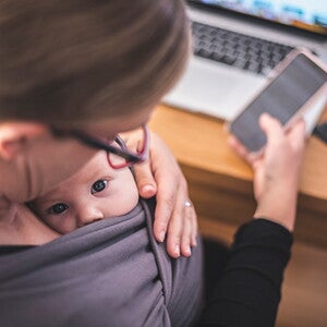 Mom looks down at baby in sling, with a phone in her hand and laptop on desk 