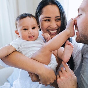 Mom holds baby, smiling. Baby has foot on dad’s nose.